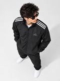 A man wearing a black Adidas track jacket with white shoulder stripes, black track pants, white sneakers, and black sunglasses, photographed from a high-angle perspective against a light gray studio background.
