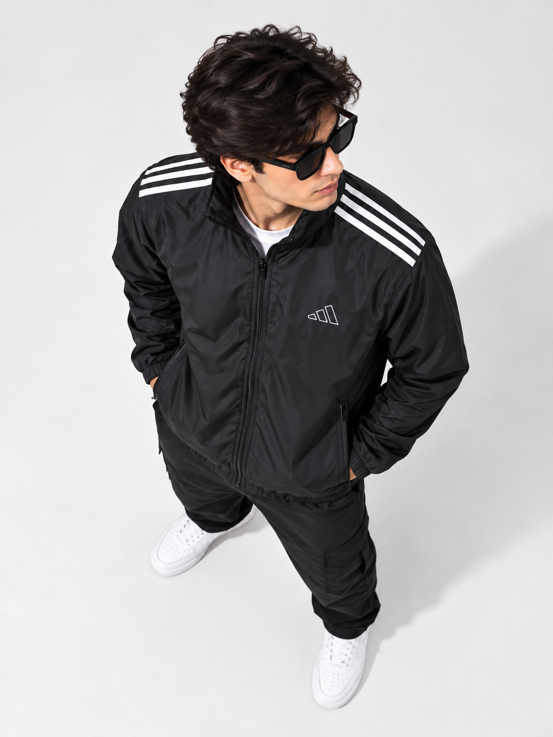 A man wearing a black Adidas track jacket with white shoulder stripes, black track pants, white sneakers, and black sunglasses, photographed from a high-angle perspective against a light gray studio background.